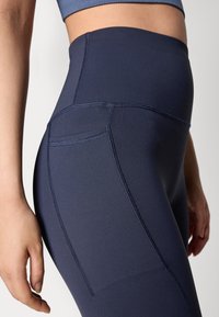 High-waisted, navy leggings made of stretchy fabric with flatlock seams and a small side pocket for storage. Smooth texture.