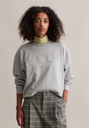 Vrouw met krullend haar, gekleed in een grijze sweatshirt met de tekst "MO'PO" en een geruite grijze broek, staat met één hand in de zak tegen een effen achtergrond.