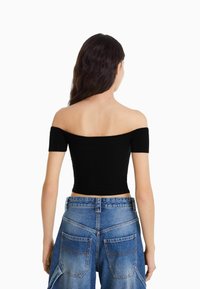 Femme aux longs cheveux noirs, portant un haut noir à épaules dénudées et un jean bleu taille haute, vue de dos sur un fond blanc.