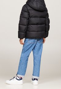 Schwarze Pufferjacke mit Kapuze, mit horizontalen Nähten und einem Patch am linken Ärmel; kombiniert mit blauen umgeschlagenen Jeans und weißen Sneakers.