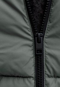 Mörkgrön vadderad jacka med svart dragkedja och texturerat innerfoder. Har quiltade mönster och en märkesetikett nära dragkedjan.