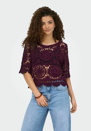 Młoda kobieta o kręconych włosach, ubrana w fioletowy szydełkowany top na czarnym podkoszulku oraz jasnoniebieskich dżinsach, stojąca z jedną ręką w kieszeni.