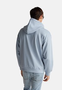 Hoodie azzurro chiaro realizzato in tessuto morbido, con un ampio cappuccio, maniche lunghe e una vestibilità casual. Cuciture visibili lungo l'orlo e i polsini.