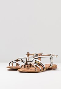 Sandalias planas de tiras en blanco y negro, que combinan materiales lisos y texturizados, con un acento dorado en la tira del tobillo.