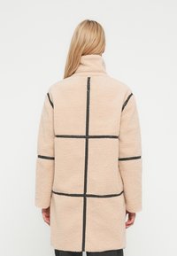 Personne blonde portant une longue veste sherpa beige avec des coutures noires horizontales et verticales, debout de dos sur un fond uni.