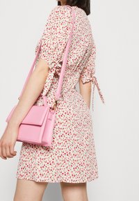 Kvinna iklädd en vit blommig klänning med rosa och röda blommor, bär en liten rosa axelväska med lång rem över axeln.