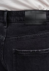 Πίσω τσέπη μαύρου τζιν με μαύρη δερμάτινη ετικέτα μάρκας ραμμένη πάνω από την τσέπη, με φόντο λευκό ύφασμα.