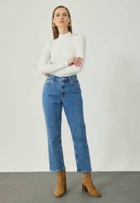 Weißes Langarmoberteil mit Schulterknöpfen, kombiniert mit hoch taillierten, hellblauen, geraden Jeans und braunen, Absatz-Stiefeletten.