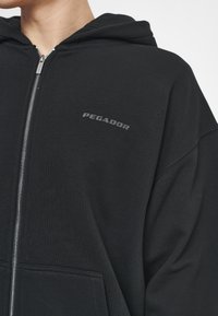Schwarze Zip-up Hoodie aus weicher Baumwolle, mit einer Fronttasche und grauem 'PEGADOR'-Logo auf der linken Seite. Glatte Textur, lockere Passform.