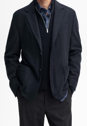 Mann trägt einen schwarzen Blazer über einem schwarzen Pullover mit Reißverschluss und einem blauen Hemd mit Kragen, mit den Händen in den Blazertaschen vor einem schlichten Hintergrund.