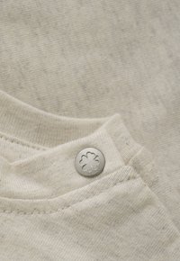 Nahaufnahme von beigem Stoff mit einem silbernen Knopf, der ein geprägtes Kleeblatt mit vier Blättern auf einer Naht des Kleidungsstücks zeigt.