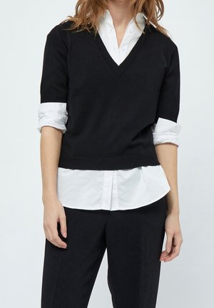 Personne portant un pull noir à manches courtes en V par-dessus une chemise blanche à boutons avec les manches retroussées et un pantalon noir, debout devant un fond uni.