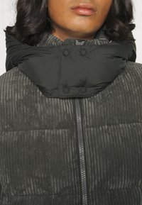 Personne portant une veste côtelée gris foncé avec un col rembourré haut et des boutons-pression noirs, fermeture éclair fermée à l'avant.