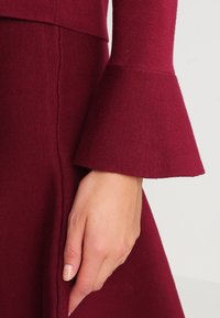 Bordowa sukienka dzianinowa z rozszerzanymi rękawami i gładką fakturą, z widocznym przeszyciem wzdłuż bocznego szwu.