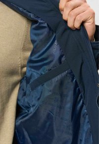 Main tirant une veste bleu marine pour révéler la doublure intérieure brillante avec le logo répété de U.S. Polo Assn. et une poche intérieure sur la poitrine.