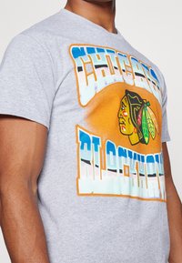 Camiseta de algodón gris con un estampado gráfico colorido que presenta el texto "Chicago Blackhawks" y un logo de cabeza nativa americana en colores vibrantes.