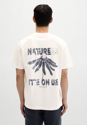 Mann trägt weißes T-Shirt mit großem schwarzem Blumenmotiv und dem Text „NATURE IT'S ON US“ auf der Rückseite, steht vor einem schlichten Hintergrund.