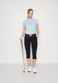 Polo de manga curta azul claro com gola zipada, calças pretas curtas e sapatos de golfe brancos. Um taco de golfe é segurado na mão direita.