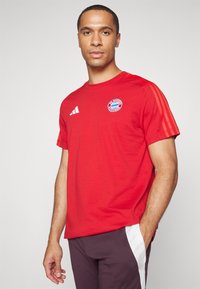 Rød atletisk t-skjorte i bomullsblanding, med hvit Adidas-logo og Bayern München-badge. Korte ermer med tre hvite striper på skuldrene.