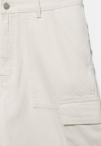 Gros plan d’un pantalon en denim écru montrant une poche avant, une passants de ceinture, un bouton et une poche à rabat avec des détails de couture.