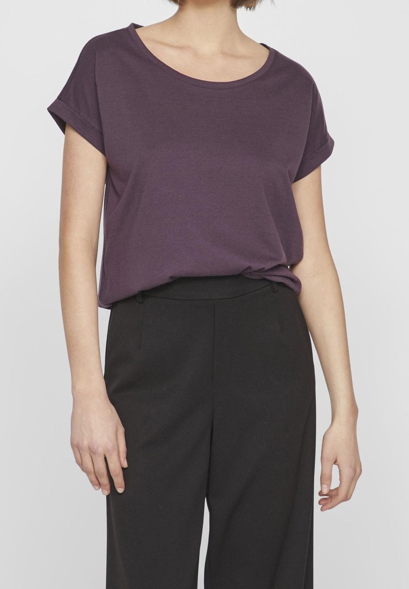 Personne portant un t-shirt violet foncé à manches courtes rentré dans un pantalon noir ample taille haute sur un fond uni.