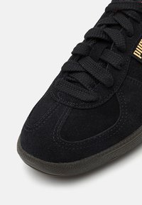 Baskets en daim noir avec des lacets noirs texturés et une semelle en caoutchouc foncé, avec un petit logo Puma doré sur le côté.