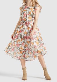 Frau in einem ärmellosen, floralen Midikleid mit Rüschen und beigen Wildleder-Ankle-Boots, die vor einem schlichten Hintergrund steht.