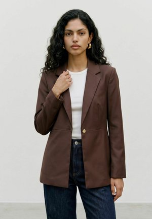 Kvinde med langt krøllet hår bærer guldfarvede øreringe, hvid top, brun blazer og mørkeblå jeans, stående mod en ensfarvet lys baggrund.