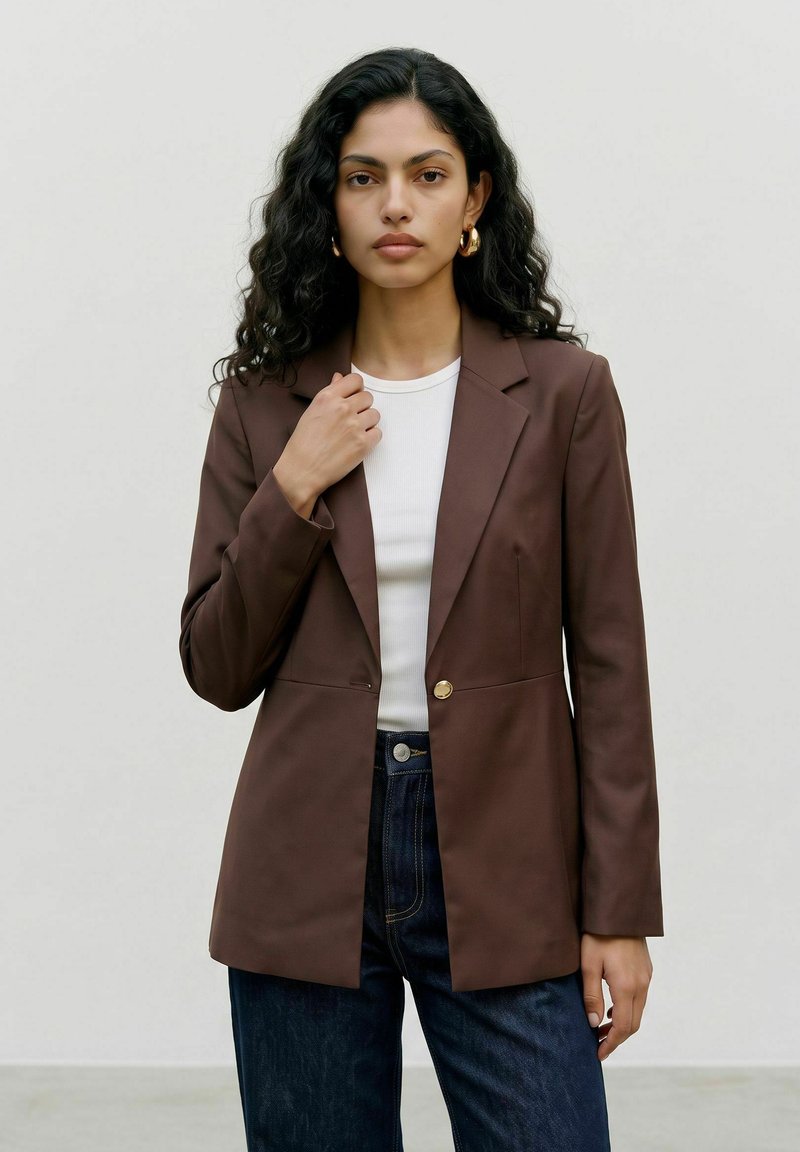 Femme aux longs cheveux bouclés porte des boucles d'oreilles créoles dorées, un haut blanc, un blazer marron et un jean bleu foncé, debout devant un fond clair uni.