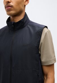 Άνδρας που φοράει μαύρο αμάνικο γιλέκο με φερμουάρ πάνω από μπεζ κοντομάνικο πουκάμισο, με το λογότυπο "BOSS" στο αριστερό στήθος.