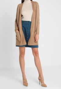 Cárdigan beige con abertura frontal y dos bolsillos, combinado con una falda de mezclilla azul con botones. Modelo lleva zapatos de tacón altos beige.