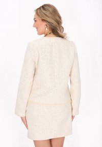 Hellbeige Tweedjacke mit langen Ärmeln, strukturierter Oberfläche und minimalistischem Schnitt. Sie hat einen geraden Saum und dezente Nähdetails.