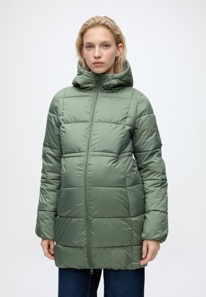 Femme portant une longue doudoune matelassée verte avec capuche, debout devant un fond clair uni.