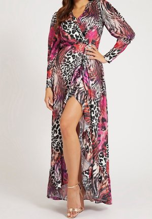 Kobieta w długiej, wielokolorowej sukience w zwierzęcy wzór z rozcięciem do uda i sandałach na wysokim obcasie w kolorze różowego złota, pozująca z ręką na biodrze.