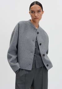 Femme avec les cheveux tirés en arrière, portant des vestes en laine grises à plusieurs couches et un pantalon gris foncé, se tenant devant un fond clair et uni.