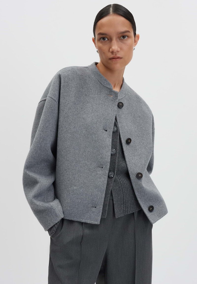 Femme avec les cheveux tirés en arrière, portant des vestes en laine grises à plusieurs couches et un pantalon gris foncé, se tenant devant un fond clair et uni.