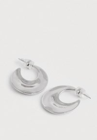 Sileda siled hõbedased poolkuu kujulised kõrvarõngad, millel on selged plastist tagused, valgel pinnal.