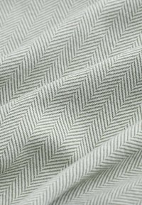 Gros plan sur un tissu gris clair avec un motif chevrons texturé et de doux plis créant des ombres et des reflets diagonaux.