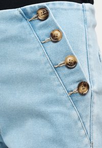 Υφασμα από ανοιχτό μπλε τζιν με τέσσερα καφέ κουμπιά, στερεωμένα διαγώνια σε ένδυμα με ψηλή μέση, που δείχνει λεπτομέρειες υφής και ραφών.