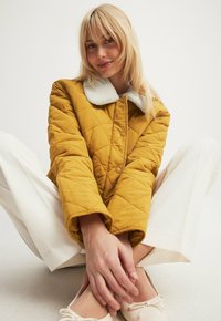 Paduotinis geltonas švarkas su minkštu baltu apykakle, su mygtukų užsegimu ir laisvu siluetu. Modelis sėdi sukryžiavęs kojas baltais džinsais.