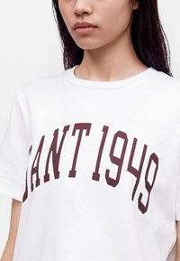 Jeune personne aux longs cheveux foncés portant un t-shirt blanc avec le texte "GANT 1949" imprimé en grandes lettres marron.