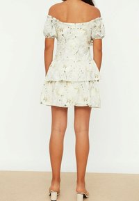 Frau trägt ein schulterfreies, kurzes Blumenkleid mit Puffärmeln und gestuftem Rock, von hinten gezeigt, steht auf einem beigen Boden.