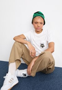 T-shirt bianca con dettaglio della tasca, pantaloni baggy color khaki e scarpe bianche con logo nero. Berretto lavorato a maglia verde e nero. Sfondo in moquette blu.