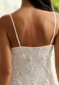 Rochie albă din material texturat, cu detalii franjurate, bretele subțiri și un decolteu neted, evidențiind un design simplu și elegant.