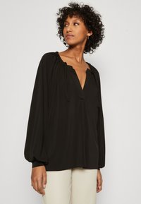Svart bluse med lange puffermer, v-hals og snorakkenter. Mykt, flytende stoff med lett struktur for ekstra detalj.