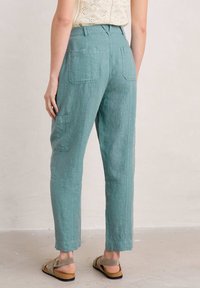Light teal linen trousers with a straight fit, featuring two back pockets and raw hem details. Sandals are worn for a casual look.