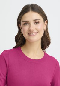 Rózsaszín kötött pulóver kerek nyakkivágással, sima textúrával. A modell apró karika fülbevalót visel, és hosszú, hullámos haja van.