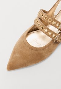 Scarpa con punta affusolata in suede color cammello con cinghie incrociate e borchie dorate. Interno liscio dorato visibile attraverso il design con ritagli.