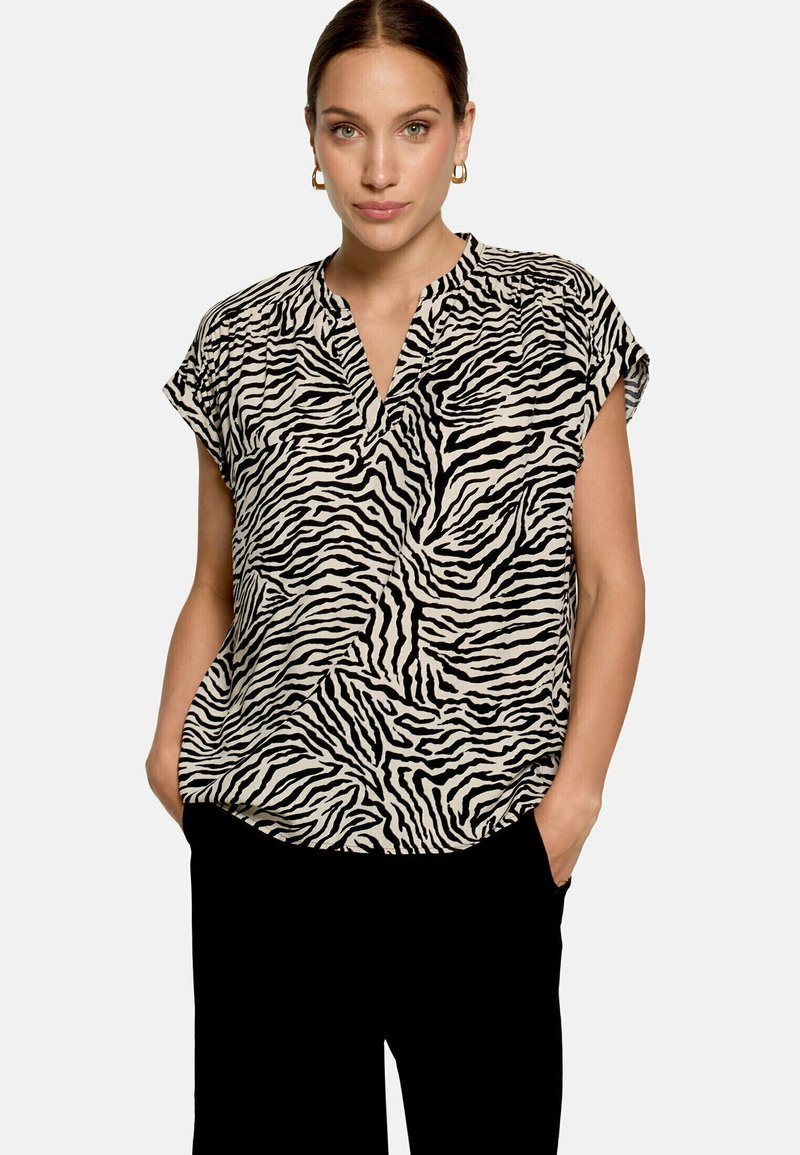Femme portant un chemisier à manches courtes imprimé zèbre noir et blanc avec col en V et pantalon noir, mains dans les poches, fond clair uni.