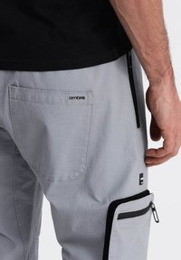 Lichtgrijze broek met een gestructureerde afwerking, voorzien van een achterzak met logo, zijzak met rits en zwarte accenten langs de rits.